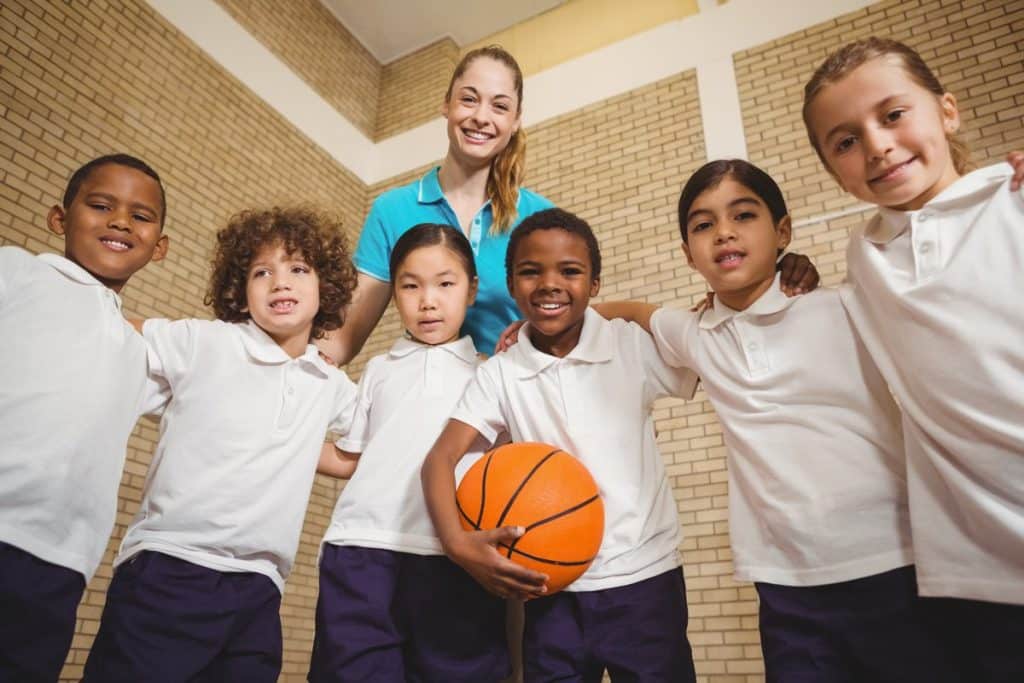 Oposiciones de Educación Física: Grupo de niños con una pelota de baloncesto y una profesora de educación física tras ellos.