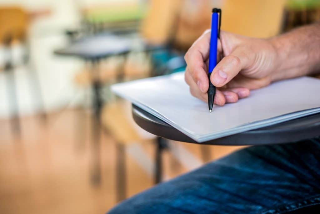 Hombre escribiendo en una hoja de papel sobre un pupitre en un aula.